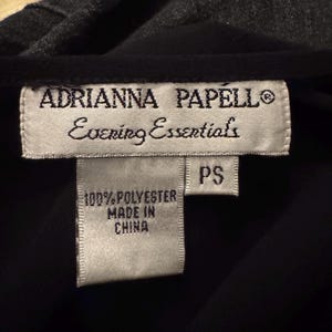 May include: Close-up of a black garment with a gray collar. The label reads "ADRIANNA PAPELL Evening Essentials" and "100% POLYESTER MADE IN CHINA". The size is "PS".