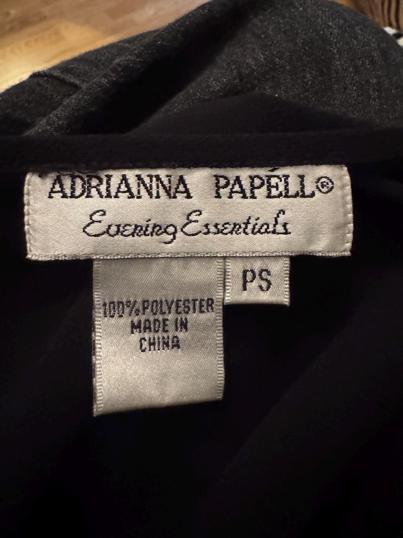 May include: Close-up of a black garment with a gray collar. The label reads "ADRIANNA PAPELL Evening Essentials" and "100% POLYESTER MADE IN CHINA". The size is "PS".