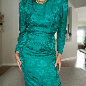 May include: A teal green dress with a floral pattern. The dress has long sleeves and a fitted silhouette. It features a round neckline and a gold chain necklace.