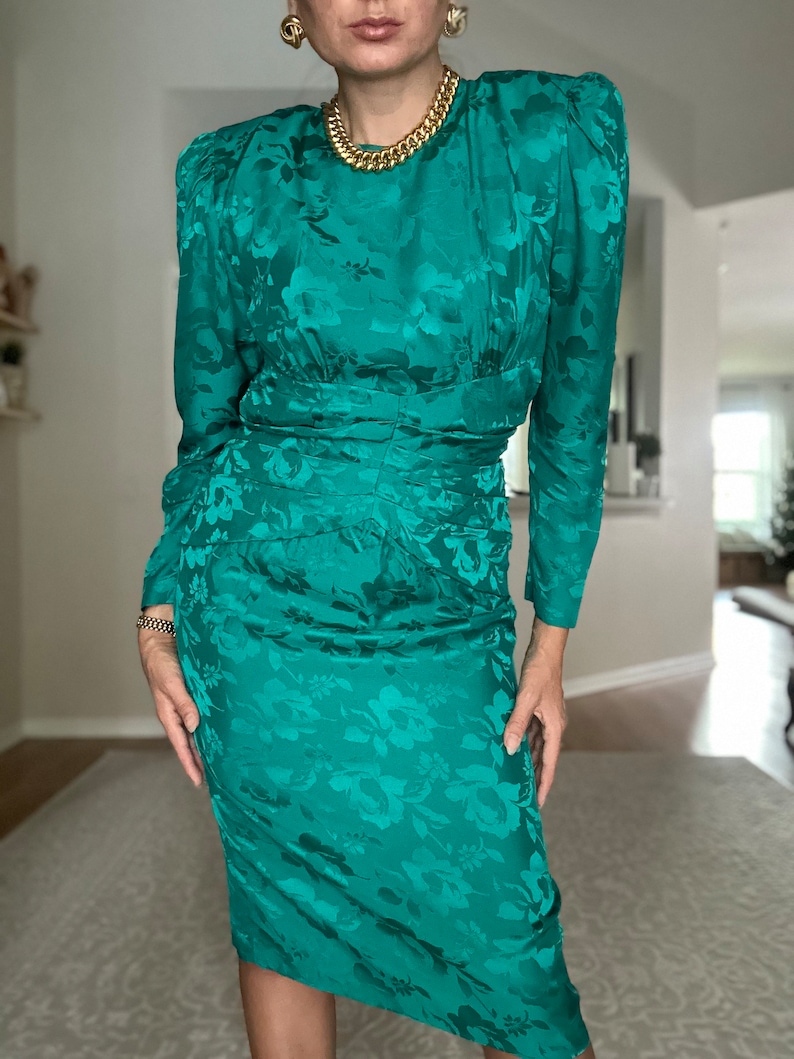 May include: A teal green dress with a floral pattern. The dress has long sleeves and a fitted silhouette. It features a round neckline and a gold chain necklace.