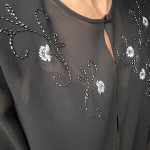 May include: Black sheer top with floral embroidery. The top features delicate white flower and beaded vine designs across the front. A small button closure is at the neckline. The fabric appears lightweight and slightly translucent.