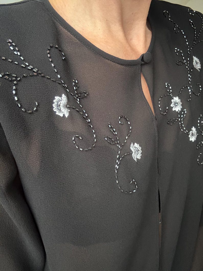 May include: Black sheer top with floral embroidery. The top features delicate white flower and beaded vine designs across the front. A small button closure is at the neckline. The fabric appears lightweight and slightly translucent.
