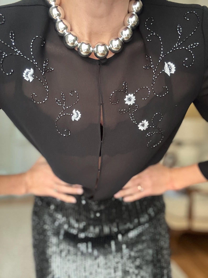 May include: Black sheer top with floral embroidery and a silver beaded necklace. The top has a split front and long sleeves. The bottom half of the image shows a black sequined skirt.