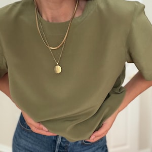 May include: An olive green short-sleeved top with a smooth finish, paired with blue denim jeans. The outfit is accessorized with a layered gold-tone necklace featuring a flat chain and a circular pendant, along with large gold-tone dome earrings.