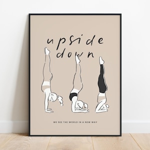 May include: A framed art print with a beige background, depicting three figures in handstand poses. The words "upside down" are written above the figures in a handwritten font. The phrase "WE SEE THE WORLD IN A NEW WAY" is written below.