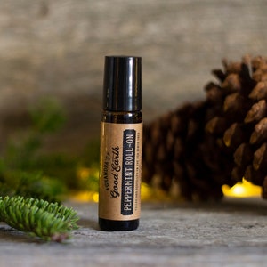May include: A brown glass roll-on bottle with a black cap. The label reads "Grandpa's Good Earth Peppermint Roll-On, Tilled and distilled in the USA since 1953." The bottle is on a wooden surface with a pine cone and greenery in the background.