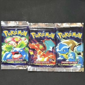 May include: Three sealed Pokémon trading card game packs. Each pack features a different Pokémon character illustration in vibrant colors. The packs are labeled "Pokémon" in yellow and blue, with the text "TRADING CARD GAME" and "11 Tradable Game Cards".