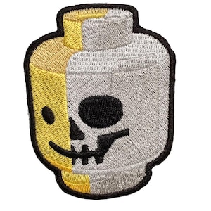 Peut inclure: Écusson brodé avec un design divisé : une moitié est jaune avec un visage souriant, l'autre un crâne gris. Le patch a une bordure noire et est en forme de tête de brique jouet.