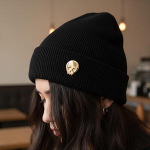 Gold minimalist human skull enamel pin on a black knitted beanie worn by a person with long, dark hair. The person is looking down, partially obscuring their face. The blurred background suggests a cafe or indoor setting, embodying a "soft goth" or "artist" aesthetic.