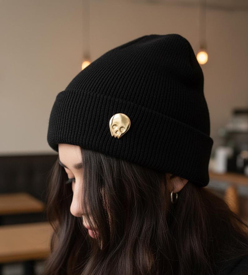Gold minimalist human skull enamel pin on a black knitted beanie worn by a person with long, dark hair. The person is looking down, partially obscuring their face. The blurred background suggests a cafe or indoor setting, embodying a "soft goth" or "artist" aesthetic.