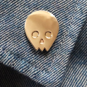 Close-up of a gold minimalist human skull enamel pin (approx. 1 inch) pinned to a denim jacket or shirt. The textured blue denim fabric and contrast stitching are visible. This shows the gold skull lapel pin as a stylish and subtle accessory for everyday wear.