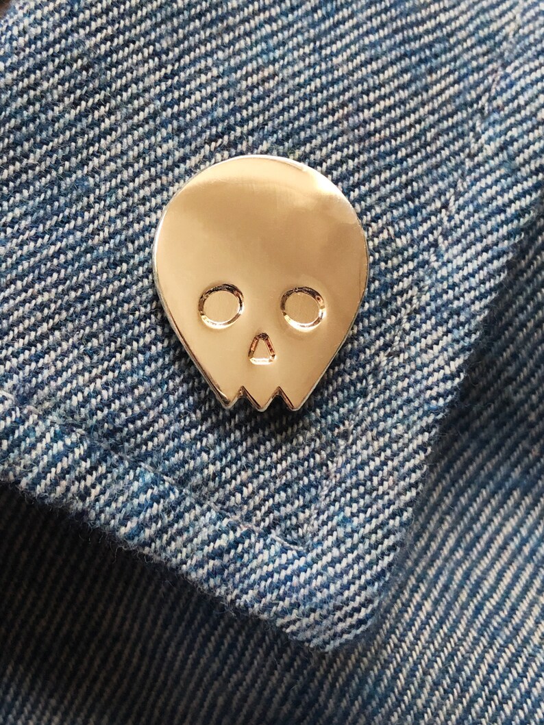 Close-up of a gold minimalist human skull enamel pin (approx. 1 inch) pinned to a denim jacket or shirt. The textured blue denim fabric and contrast stitching are visible. This shows the gold skull lapel pin as a stylish and subtle accessory for everyday wear.