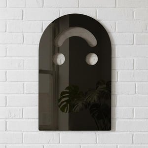 Dark (black) smiley face acrylic mirror, 17.5" tall, hanging on a white brick wall. It reflects a window on the left and a large, dark green monstera plant on the right. The moody, subdued mirror has silvery undertones, perfect for goth room decor or alternative style.