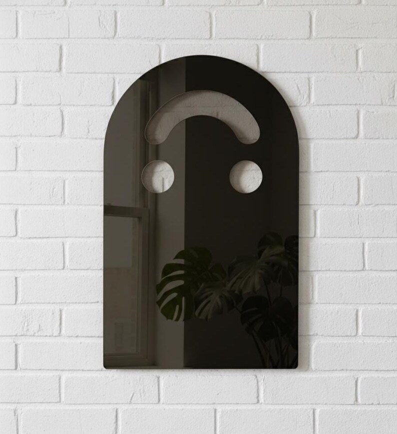 Dark (black) smiley face acrylic mirror, 17.5" tall, hanging on a white brick wall. It reflects a window on the left and a large, dark green monstera plant on the right. The moody, subdued mirror has silvery undertones, perfect for goth room decor or alternative style.