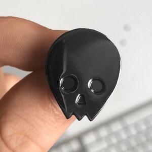 Close-up of a black minimalist human skull enamel pin (approx. 1 inch) held between a person's fingers. The dark, glossy finish of the pin is in focus, with a blurred white background. This image provides a clear view of the black alien skull pin's design and scale.