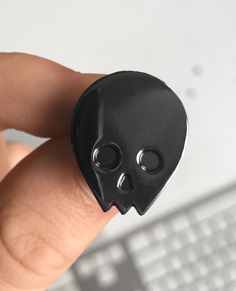 Close-up of a black minimalist human skull enamel pin (approx. 1 inch) held between a person's fingers. The dark, glossy finish of the pin is in focus, with a blurred white background. This image provides a clear view of the black alien skull pin's design and scale.