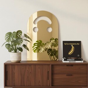 Gold smiley face acrylic mirror, 17.5" tall, standing on a dark wood mid-century modern credenza. It reflects a large green monstera plant. Next to it are three books, including "BASQUIAT," and a black vinyl record with a yellow banana, showcasing its use as unique aesthetic home decor in a stylish room.