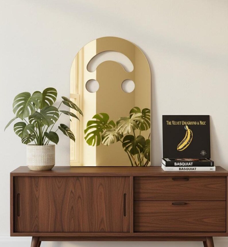 Gold smiley face acrylic mirror, 17.5" tall, standing on a dark wood mid-century modern credenza. It reflects a large green monstera plant. Next to it are three books, including "BASQUIAT," and a black vinyl record with a yellow banana, showcasing its use as unique aesthetic home decor in a stylish room.