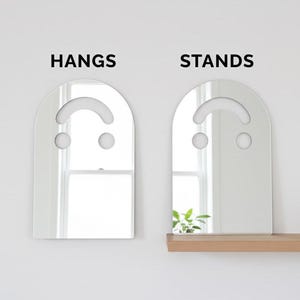 Side-by-side comparison of the silver smiley face acrylic mirror, 17.5" tall. The left mirror hangs on a white wall, labeled "HANGS." The right mirror stands on a wooden shelf, labeled "STANDS," reflecting a plant. This image highlights the versatile hanging or standing display options for this aesthetic mirror.