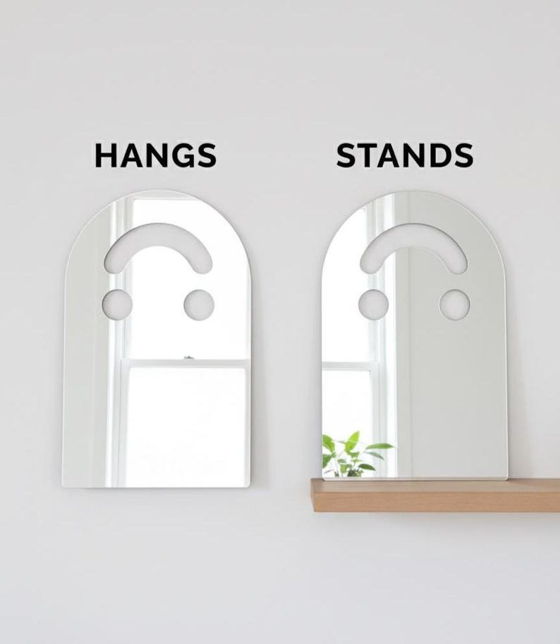 Side-by-side comparison of the silver smiley face acrylic mirror, 17.5" tall. The left mirror hangs on a white wall, labeled "HANGS." The right mirror stands on a wooden shelf, labeled "STANDS," reflecting a plant. This image highlights the versatile hanging or standing display options for this aesthetic mirror.