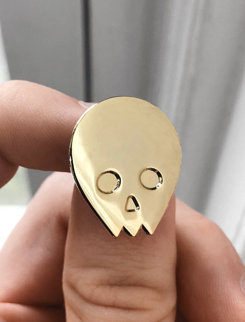 Close-up of a gold minimalist human skull enamel pin (approx. 1 inch) held between a person's fingers. The shiny, metallic gold finish of the pin is in focus, with a blurred light background. This image offers a detailed view of the gold alien skull pin's design and scale.