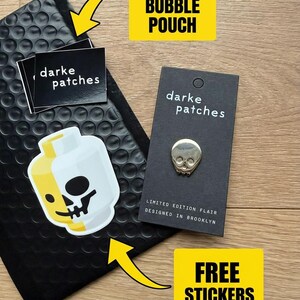 A flat layout of the gold skull pin on a black minimalist backing card, next to a black bubble pouch and some stickers. There are text callouts that say "Ships in a bubble pouch" and "Free stickers."