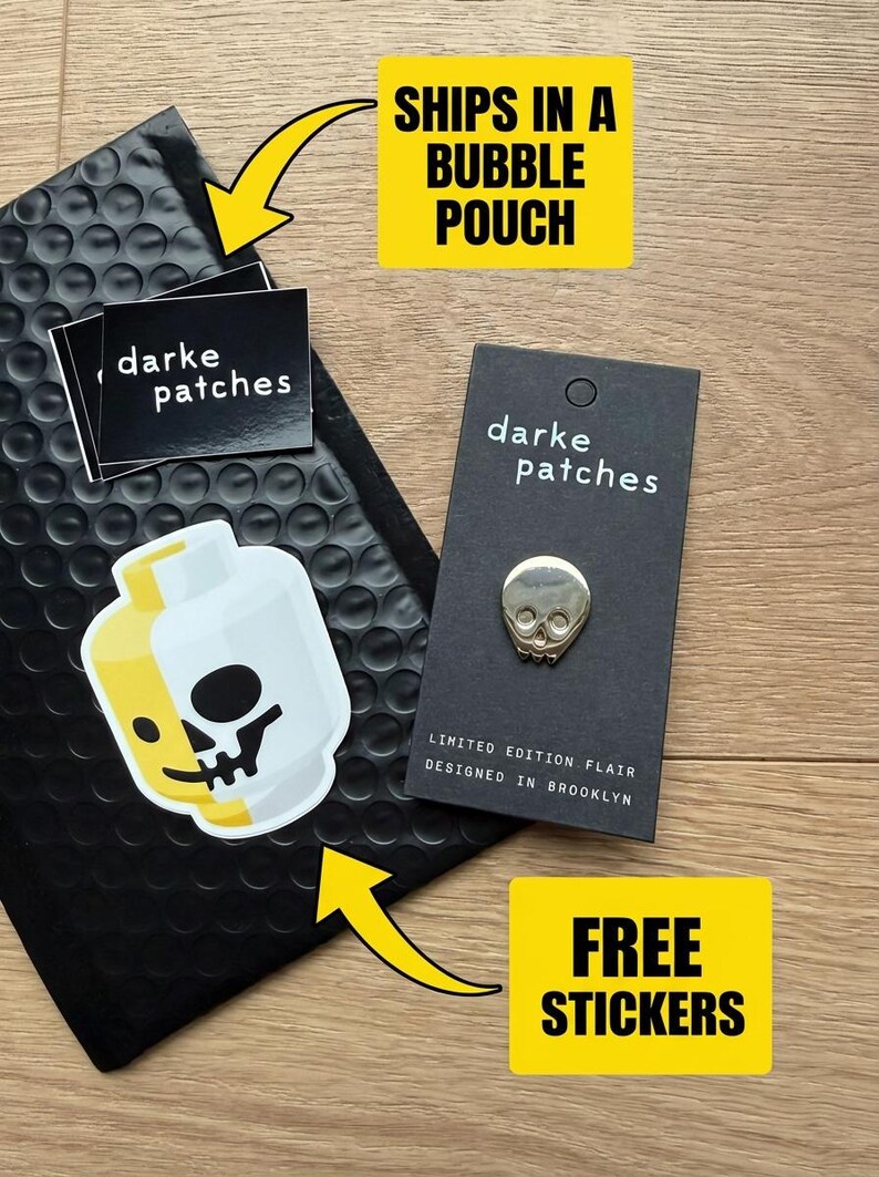 A flat layout of the gold skull pin on a black minimalist backing card, next to a black bubble pouch and some stickers. There are text callouts that say "Ships in a bubble pouch" and "Free stickers."