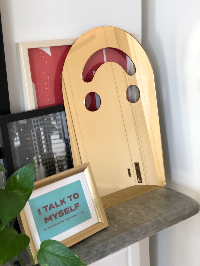 Gold smiley face acrylic mirror, 17.5" tall, standing on a grey stone shelf. It reflects red and black framed art behind it, alongside a small framed print that says "I TALK TO MYSELF" and a green plant. This funky wall art embodies aesthetic bedroom decor.