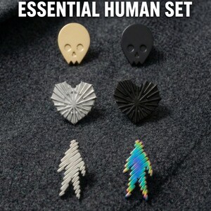 A grid of minimalist metal enamel pins on dark fabric with text above them that says "Part of the Essential Human" in all caps. At the top there is a gold skull and a black skull, in the middle there is a silver heart and a black heart, and at the bottom there is a silver soul and a rainbow soul.