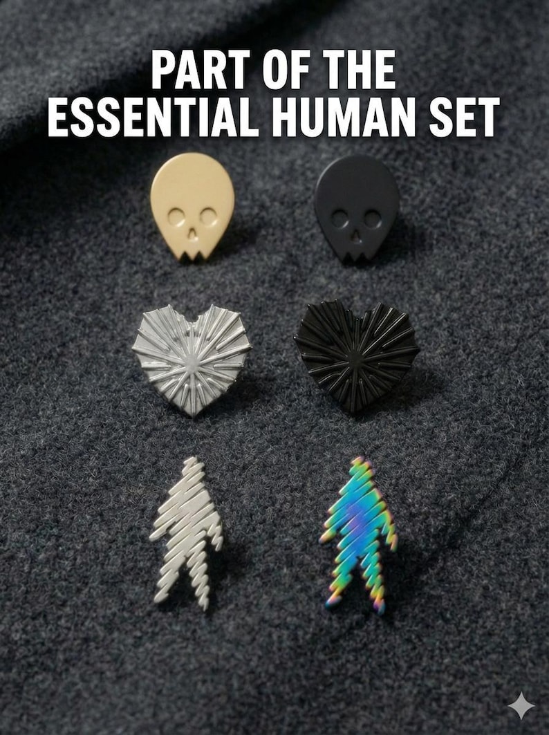 A grid of minimalist metal enamel pins on dark fabric with text above them that says "Part of the Essential Human" in all caps. At the top there is a gold skull and a black skull, in the middle there is a silver heart and a black heart, and at the bottom there is a silver soul and a rainbow soul.