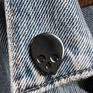 Close-up of a black minimalist human skull enamel pin (approx. 1 inch) pinned to a denim jacket or shirt. The textured blue denim fabric and contrast stitching are visible. This showcases the black skull lapel pin as a versatile "goth" or "minimalist" accessory.