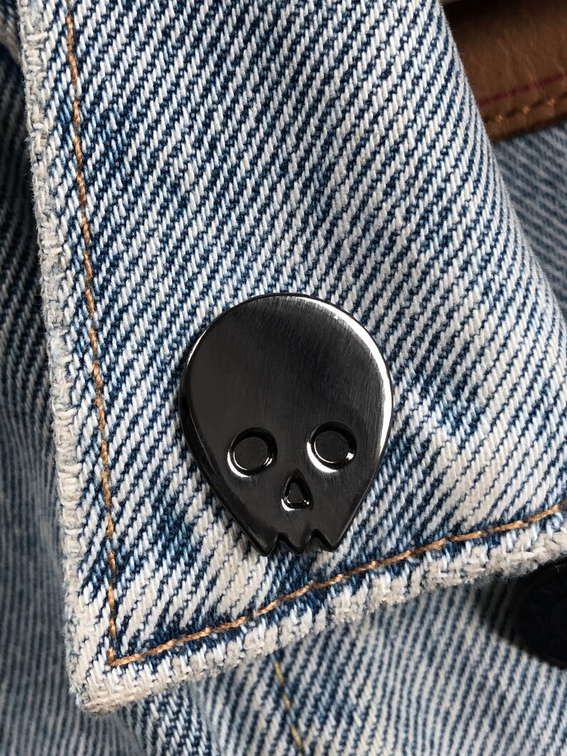 Close-up of a black minimalist human skull enamel pin (approx. 1 inch) pinned to a denim jacket or shirt. The textured blue denim fabric and contrast stitching are visible. This showcases the black skull lapel pin as a versatile "goth" or "minimalist" accessory.