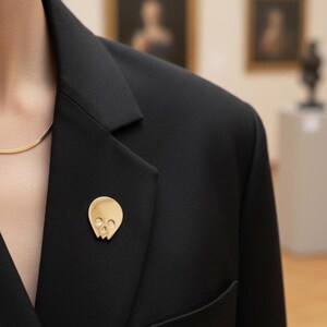 Gold minimalist human skull enamel pin on the lapel of a chic black blazer, worn by a person with a delicate gold chain necklace. The background is a softly blurred art gallery interior with paintings, showcasing the pin's "refined goth" and "artist" aesthetic.