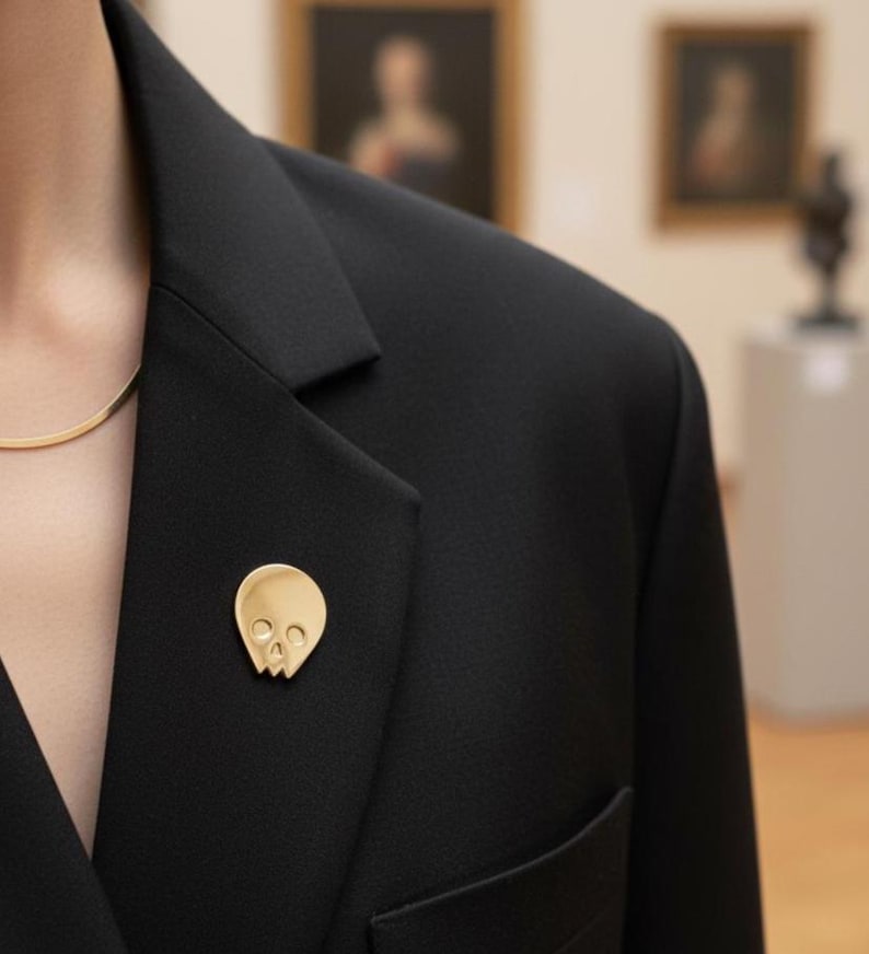 Gold minimalist human skull enamel pin on the lapel of a chic black blazer, worn by a person with a delicate gold chain necklace. The background is a softly blurred art gallery interior with paintings, showcasing the pin's "refined goth" and "artist" aesthetic.