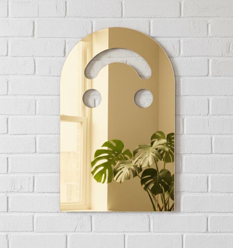 Gold smiley face acrylic mirror, 17.5" tall, hanging on a white brick wall. It reflects a bright window on the left and a large green monstera plant on the right. The warm, rich reflection offers a physical filter, ideal for funky wall art and aesthetic bedroom decor.