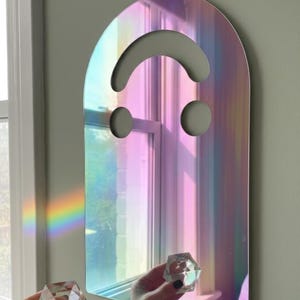Holo (rainbow) smiley face acrylic mirror, 17.5" tall, hanging on a sage green wall. A hand with black nail polish holds a clear prism, casting a rainbow onto the wall beside the mirror. The iridescent surface shimmers with pinks and blues, highlighting its ethereal quality as unique aesthetic wall art.
