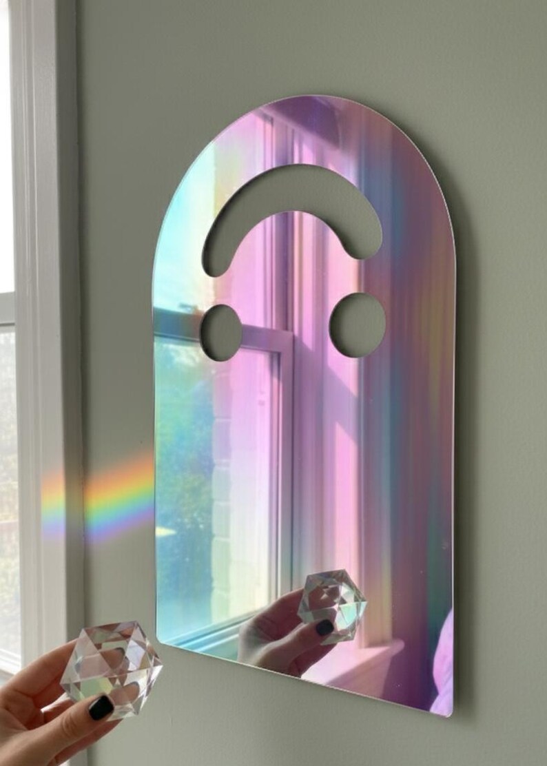 Holo (rainbow) smiley face acrylic mirror, 17.5" tall, hanging on a sage green wall. A hand with black nail polish holds a clear prism, casting a rainbow onto the wall beside the mirror. The iridescent surface shimmers with pinks and blues, highlighting its ethereal quality as unique aesthetic wall art.