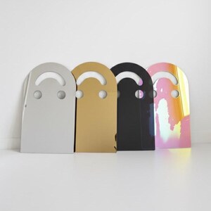 Four smiley face acrylic mirrors, 17.5" tall, leaning against a white wall in a row. From left to right: Silver, Gold, Dark (black), and Holo (rainbow). This image clearly showcases the different color options for this unique aesthetic mirror art.