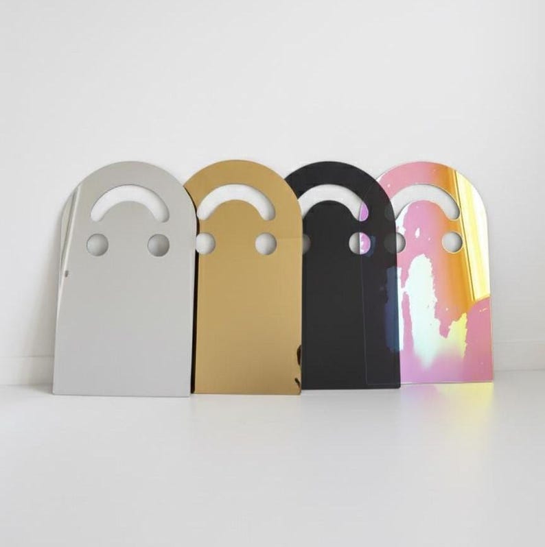 Four smiley face acrylic mirrors, 17.5" tall, leaning against a white wall in a row. From left to right: Silver, Gold, Dark (black), and Holo (rainbow). This image clearly showcases the different color options for this unique aesthetic mirror art.