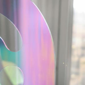 Extreme close-up of the iridescent Holo (rainbow) smiley face acrylic mirror. The focus is on the curved top edge and one of the round eye cut-outs, showing the precise laser cut and shimmering color shifts. This unique wall decor piece is shatter-resistant acrylic, 17.5" tall.