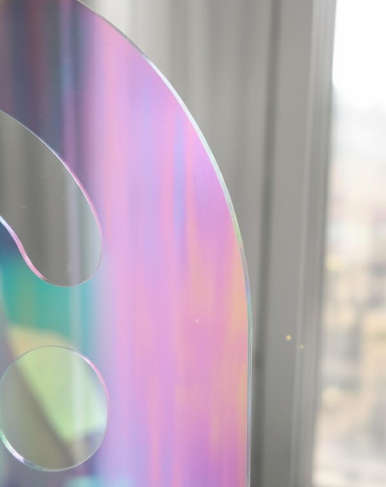 Extreme close-up of the iridescent Holo (rainbow) smiley face acrylic mirror. The focus is on the curved top edge and one of the round eye cut-outs, showing the precise laser cut and shimmering color shifts. This unique wall decor piece is shatter-resistant acrylic, 17.5" tall.
