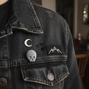 Gray minimalist human skull enamel pin and a white crescent moon pin on the lapel of a dark denim jacket. The jacket is distressed, and other small black pins and a mountain patch are visible. The background is a softly blurred, warm-toned interior, showing the pin's "soft goth" and "alternative" aesthetic.
