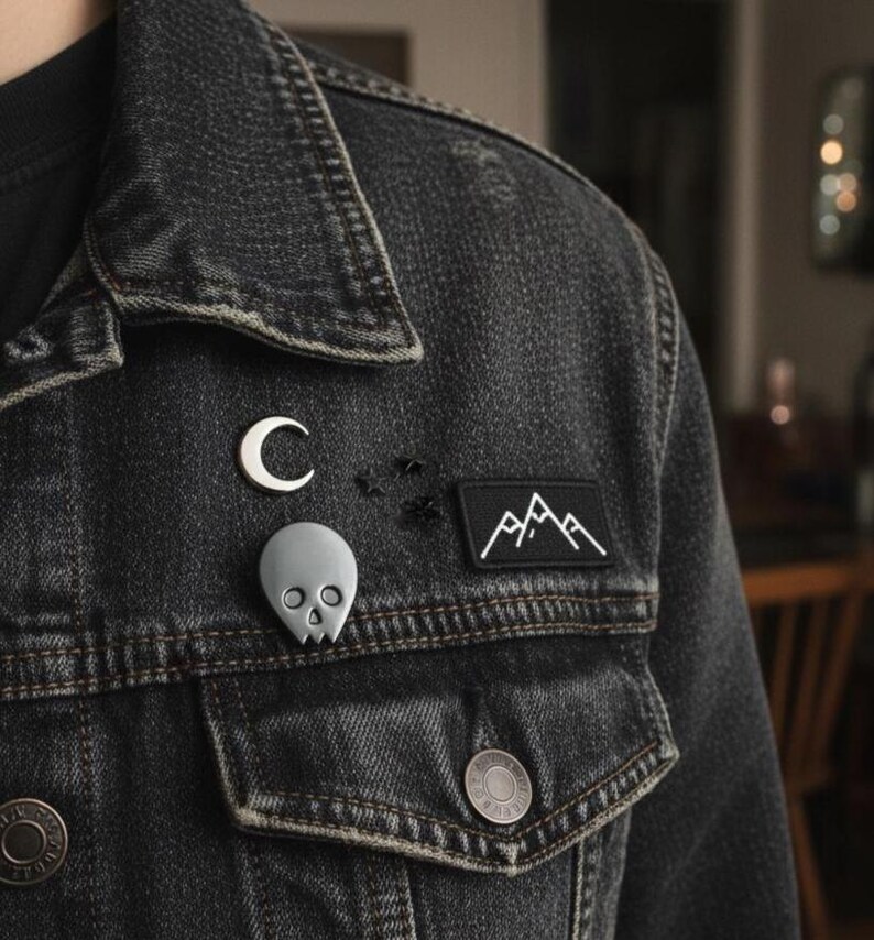 Gray minimalist human skull enamel pin and a white crescent moon pin on the lapel of a dark denim jacket. The jacket is distressed, and other small black pins and a mountain patch are visible. The background is a softly blurred, warm-toned interior, showing the pin's "soft goth" and "alternative" aesthetic.