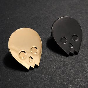 Close-up of two minimalist human skull enamel pins. The gold skull pin is on the left, and the black skull pin is on the right, both resting on a dark, textured surface. This image highlights the distinct finishes of the gold and black alien-shaped skull pins.