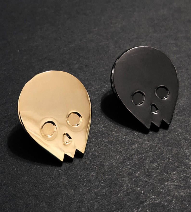 Close-up of two minimalist human skull enamel pins. The gold skull pin is on the left, and the black skull pin is on the right, both resting on a dark, textured surface. This image highlights the distinct finishes of the gold and black alien-shaped skull pins.