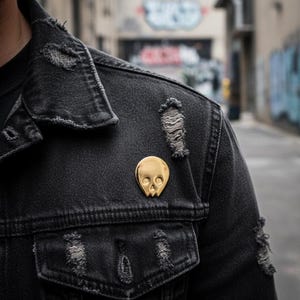 Gold minimalist human skull enamel pin on the distressed lapel of a dark denim jacket. The jacket is ripped in several places, and the background shows a blurred, graffiti-covered urban alleyway. This image captures the "goth," "punk," and "urban explorer" vibe for the gold skull pin.
