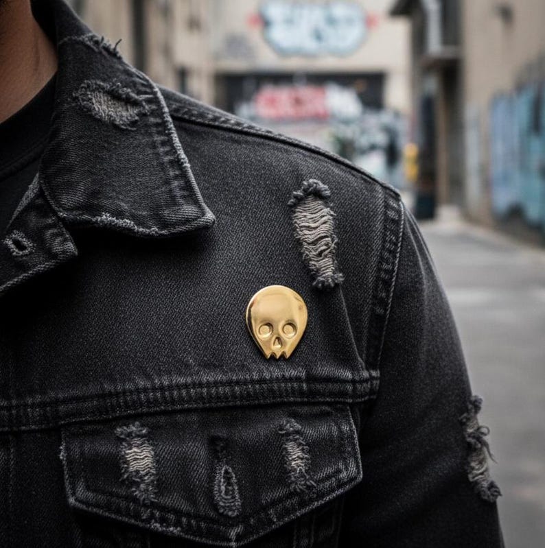 Gold minimalist human skull enamel pin on the distressed lapel of a dark denim jacket. The jacket is ripped in several places, and the background shows a blurred, graffiti-covered urban alleyway. This image captures the "goth," "punk," and "urban explorer" vibe for the gold skull pin.