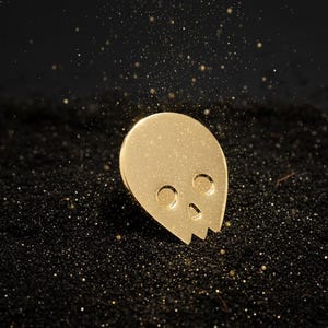 Gold minimalist human skull enamel pin (approximately 1 inch tall) sitting on a surface of fine black sand. Shimmering golden glitter or dust falls from above, creating a mystical and radiant effect around the pin. This hero shot emphasizes the "mind's power" and "radiant aura" of the gold skull.