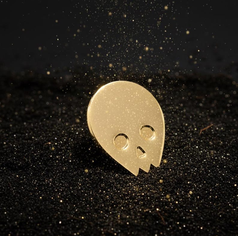 Gold minimalist human skull enamel pin (approximately 1 inch tall) sitting on a surface of fine black sand. Shimmering golden glitter or dust falls from above, creating a mystical and radiant effect around the pin. This hero shot emphasizes the "mind's power" and "radiant aura" of the gold skull.