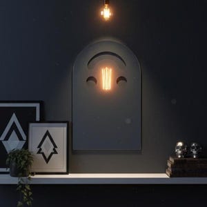 Dark (black) smiley face acrylic mirror, 17.5" tall, hanging on a dark wall above a white floating shelf. A glowing Edison bulb is reflected in the mirror's cut-out. The shelf holds two framed prints with geometric designs, a green plant, and two silver skulls on books. Perfect for gothic bedroom decor.